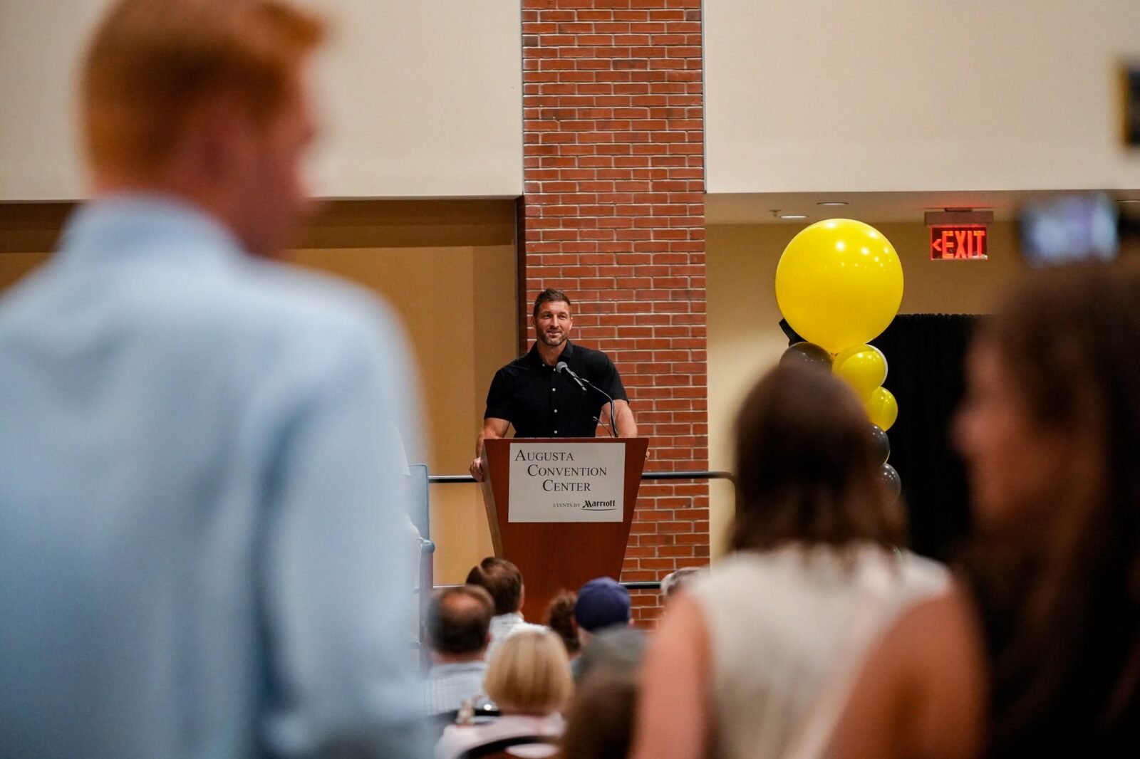 Speaker at a podium at the Augusta Convention Center with gold balloons