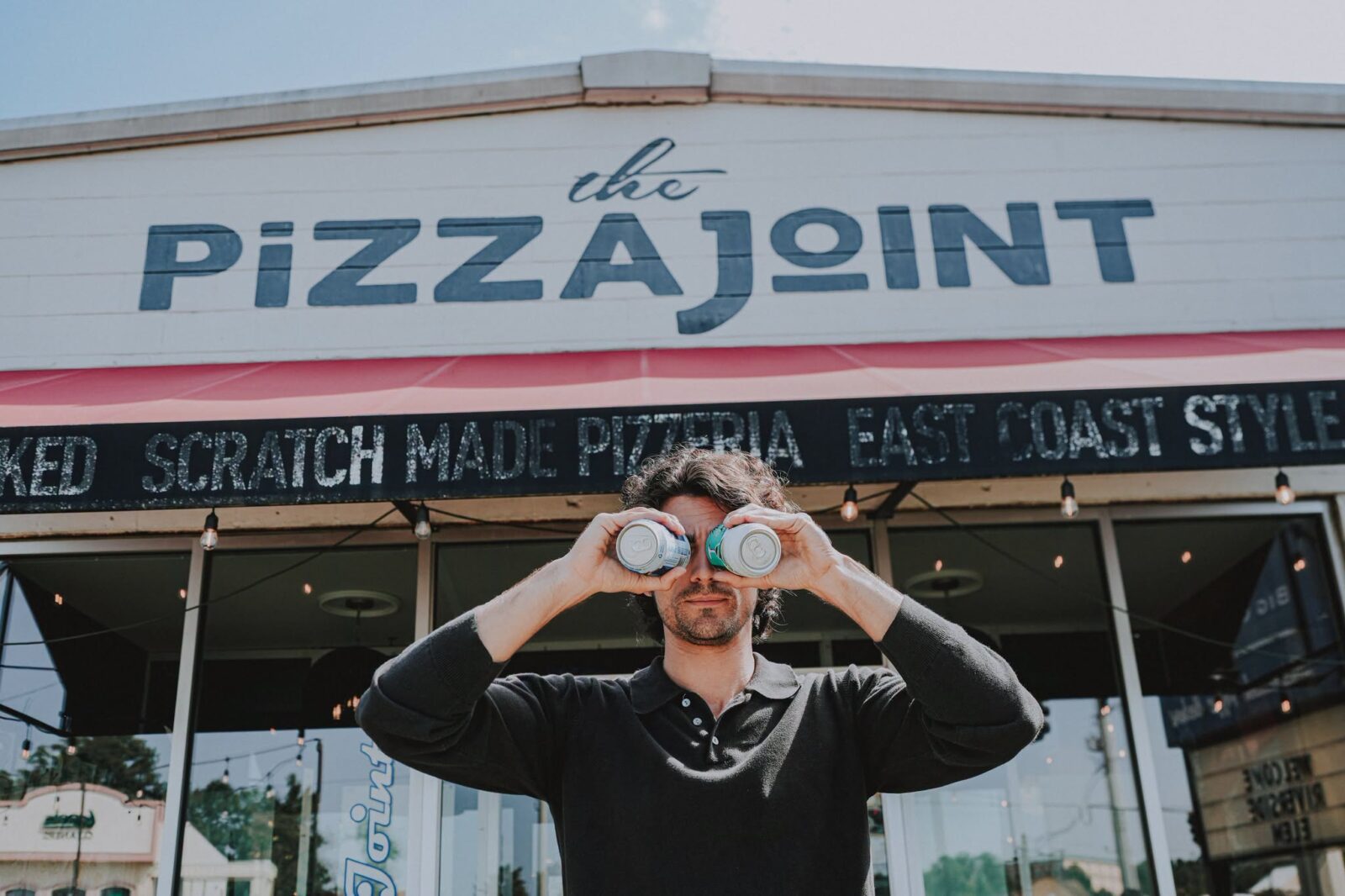 Man holding two drinks up like binoculars in front of The Pizza Joint storefront