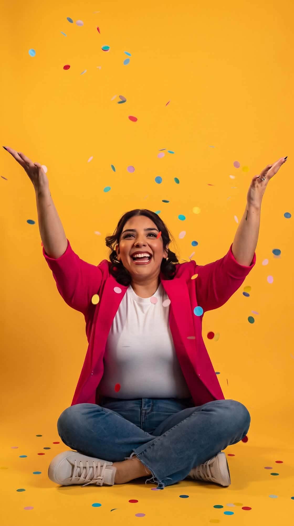 Woman in a pink blazer tossing confetti on a bright yellow backdrop in the studio